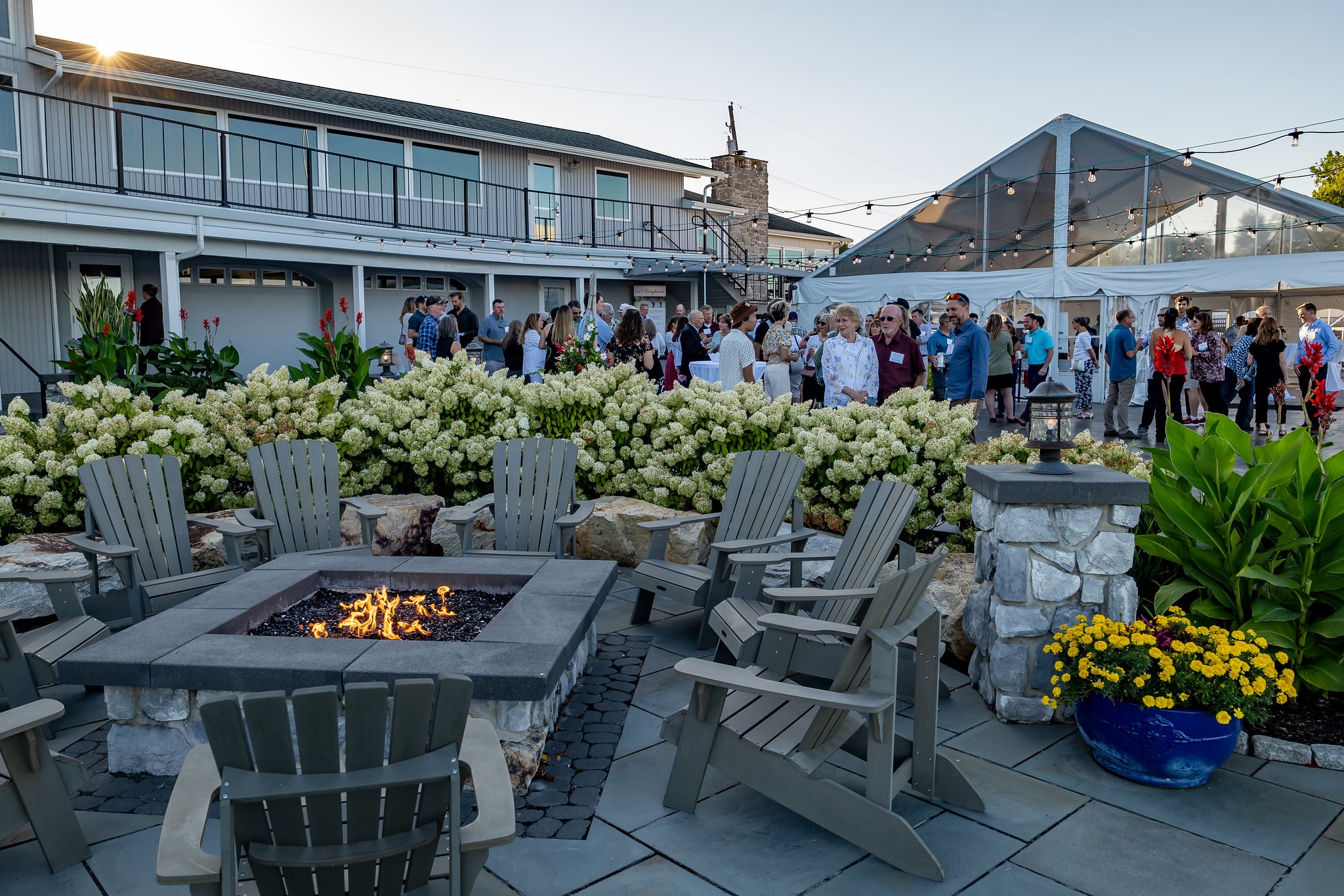 The sun sets behind River's Edge event venue with a fireplace and blooming, white hydrangeas.