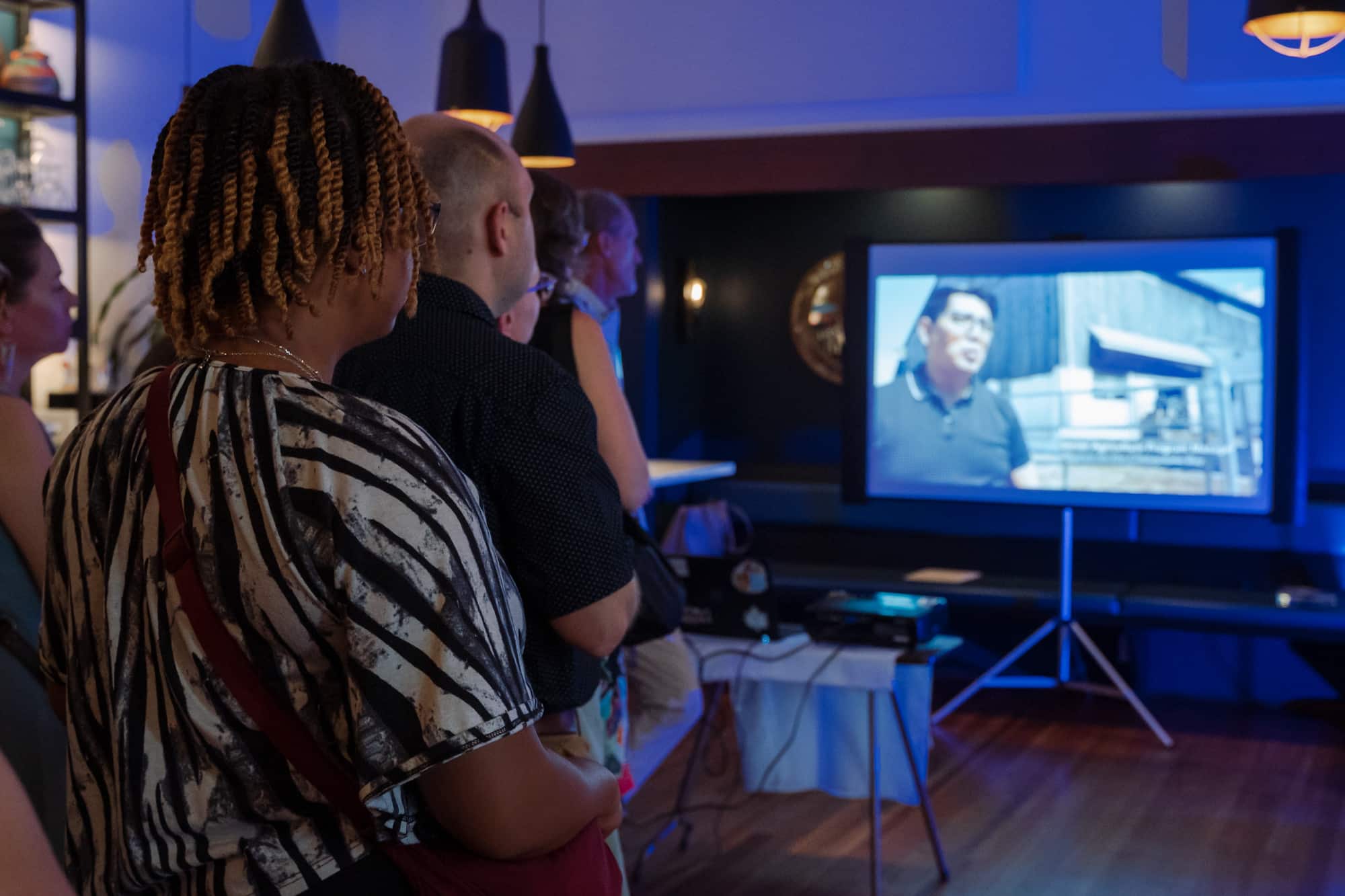 A group of people gathering around a large screen showing the Alliance's 2023 Impact Video at the Taste event.