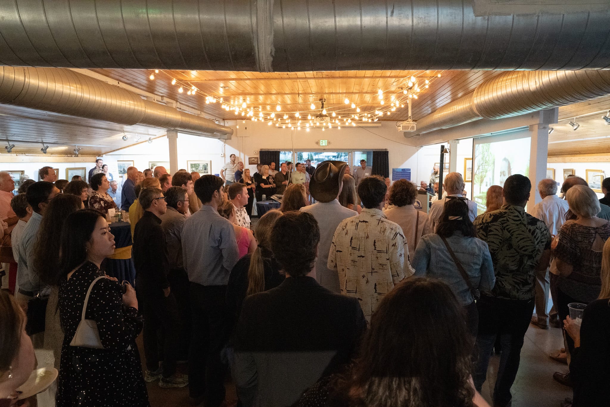 A large group of people standing under bistro lights and gathering around a large screen showing the Alliance's 2023 Impact Video at the Taste event.
