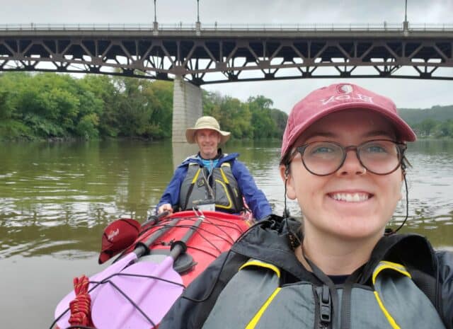 Two people floating down a river in kayaks, past a bridge.