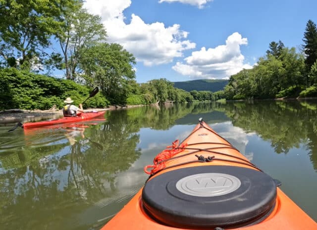 The view from inside of a kayak on a rive, with another kayaker to the left. Green trees in the foreground and sunny blue skies in the background.