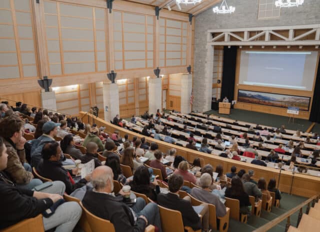 An auditorium full of people, listening to a speaker in the distance