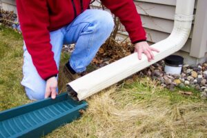 A person kneels outside a house with gray siding as they connect their downspout to a drain tray.