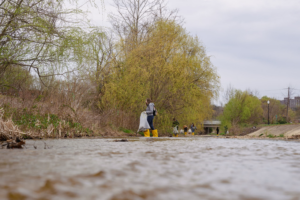 A person wearing yellow rain boots walking across a small stream picking up trash.