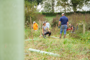 Two people, one kneeling by a tree and one standing holding a stake pounder, talk at a volunteer tree planting event on a farm.