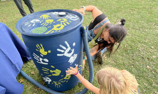 Two people kneeling and pressing one hand each to a rain barrel to imprint their handprint on the rain barrel.