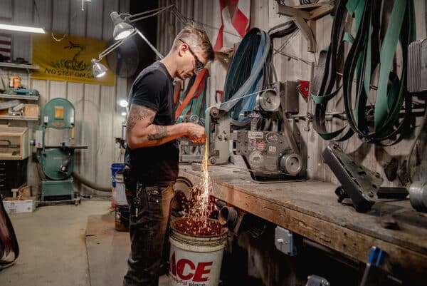 A person wearing safety goggles standing in a workshop forging steel into an oyster knife. Sparks fly below their hands and the equipment.