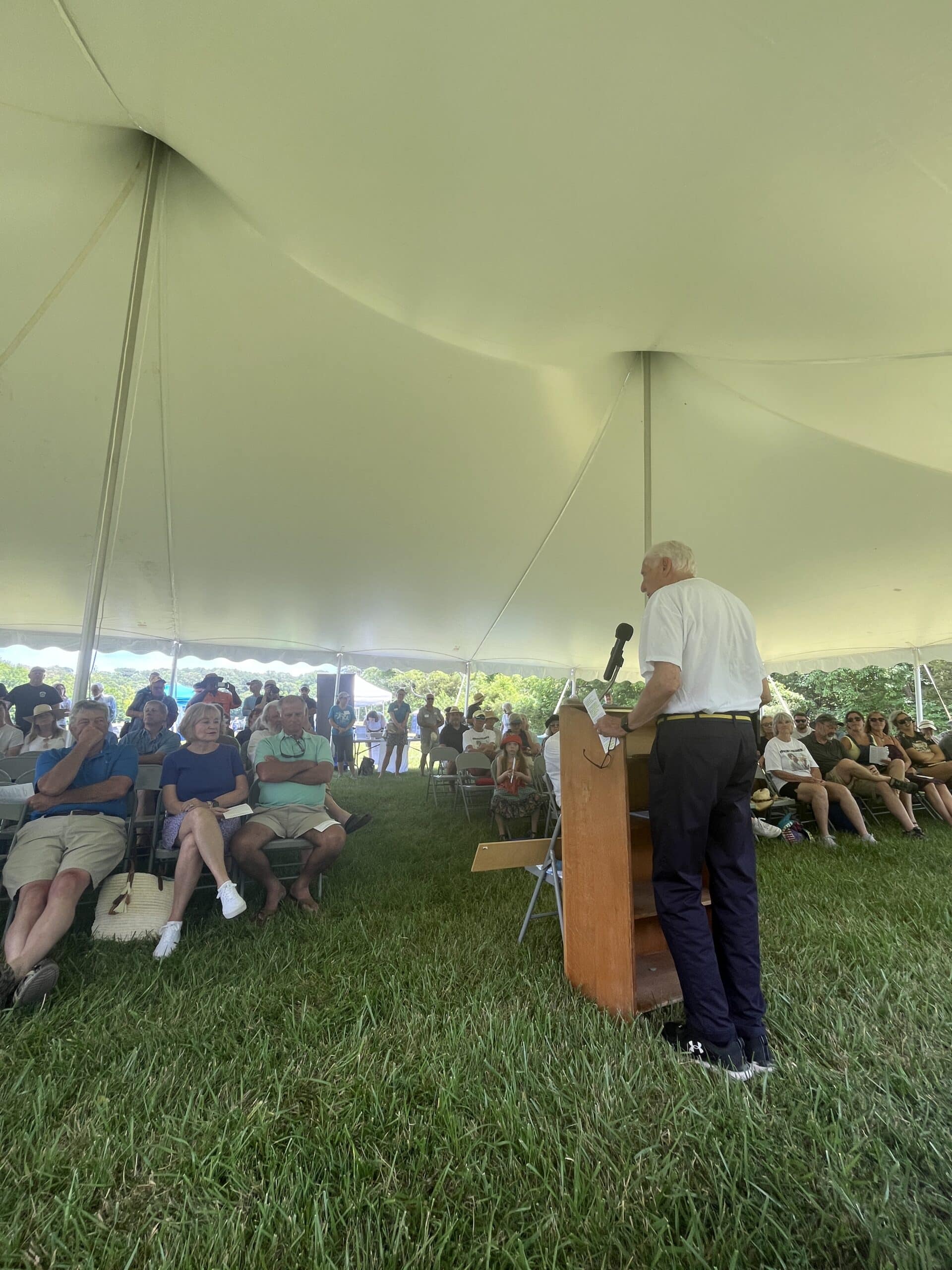 A person speaks at a podium in front of a group of people under a canopy