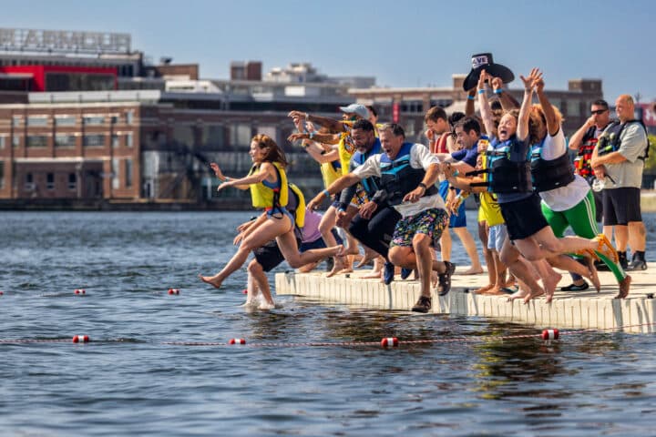 A group of people in floatation vest jump into the water off of a dock