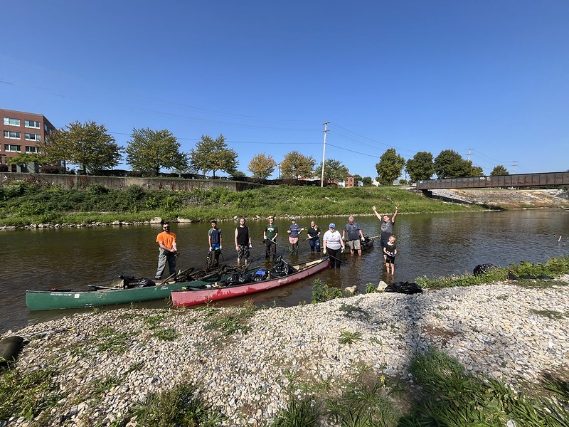 A group of volunteers stands in knee-high water in the Codorus Creek posing behind two canoes that hold trash they collected.