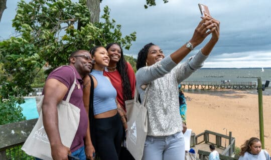 A group of people smile while one person takes a selfie with their phone