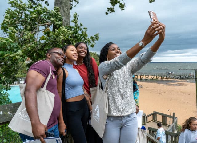 A group of people smile while one person takes a selfie with their phone