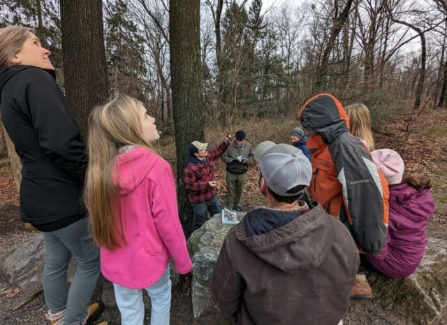 A group of people gather around a tree as someone speaks