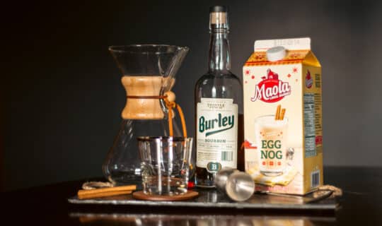 A decanter, bottle of bourbon, eggnog, and cocktail supplies sitting on a slate cutting board