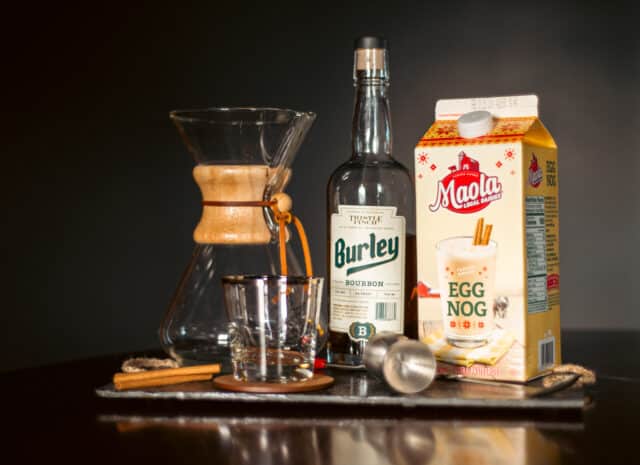 A decanter, bottle of bourbon, eggnog, and cocktail supplies sitting on a slate cutting board