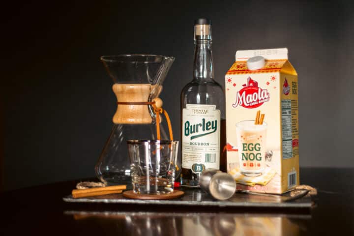 A decanter, bottle of bourbon, eggnog, and cocktail supplies sitting on a slate cutting board