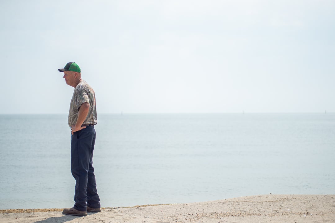 A person stands along a shoreline on a sunny day with the water in the background