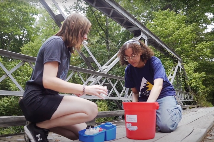 Two people on a bridge collecting water samples out of a red bucket