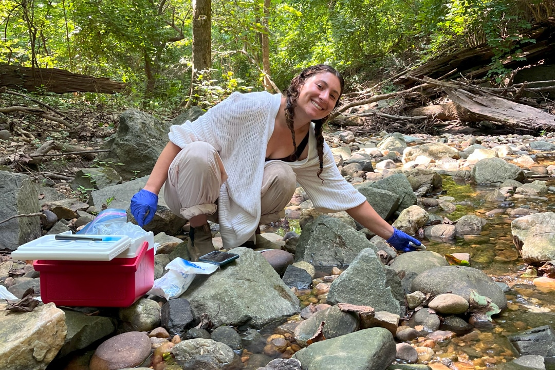 A person smiling while crouched and reaching into a creek bed.